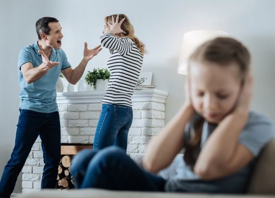 Unhappy family. Unsmiling fair-haired little girl closing her ears and sitting on the sofa while her parents shouting at each other