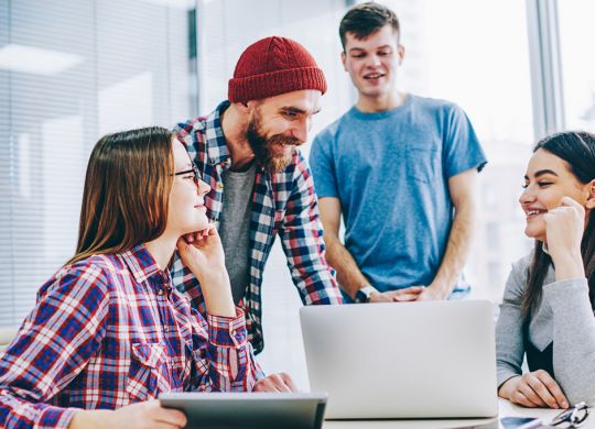 Successful group of young professionals in casual wear laughing during cooperation on design project sitting at laptop in office.Positive students teamworking on common task during brainstorming